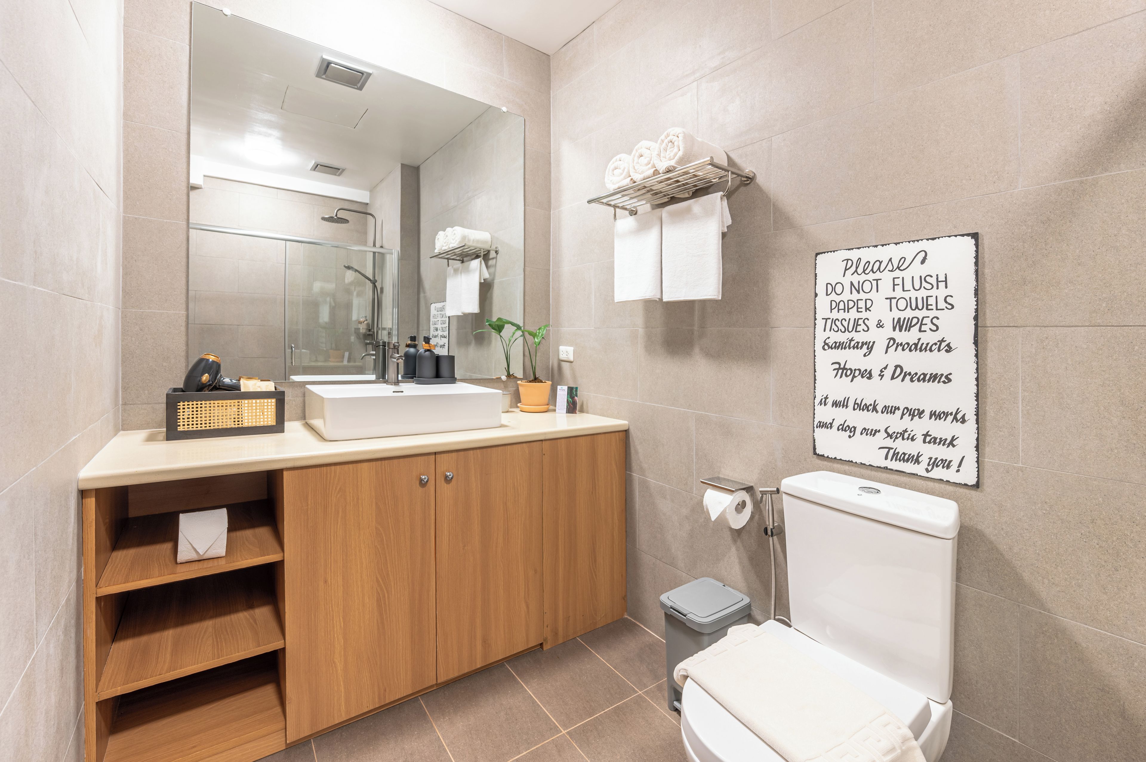 A modern bathroom with a large mirror, a sink, a toilet, and a trash bin. There is a sign on the wall with a message about not flushing paper towels.