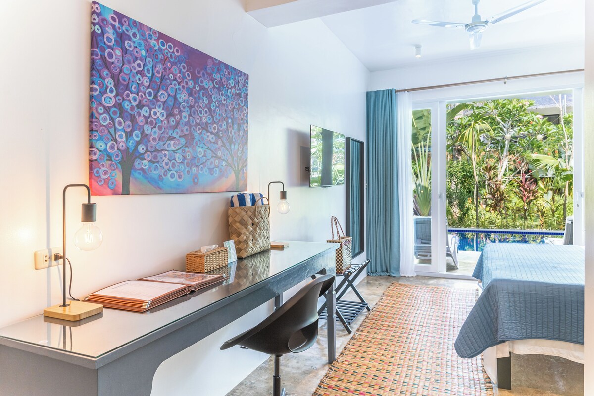 Habitación de hotel moderna con un escritorio de cristal, un cuadro abstracto grande, una cama con colcha azul y vistas a un jardín desde el balcón.