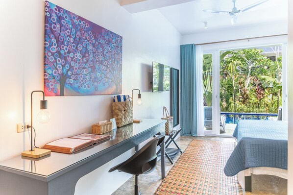A modern hotel room with a glass desk, a large abstract painting, a bed with a blue quilt, and a view of a garden through the balcony.