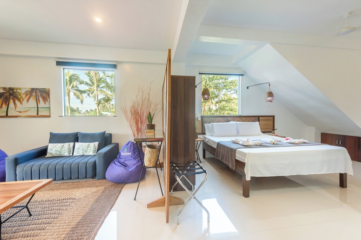 Un dormitorio moderno con cama, sofá, mesa de comedor y vistas a palmeras a través de las ventanas.