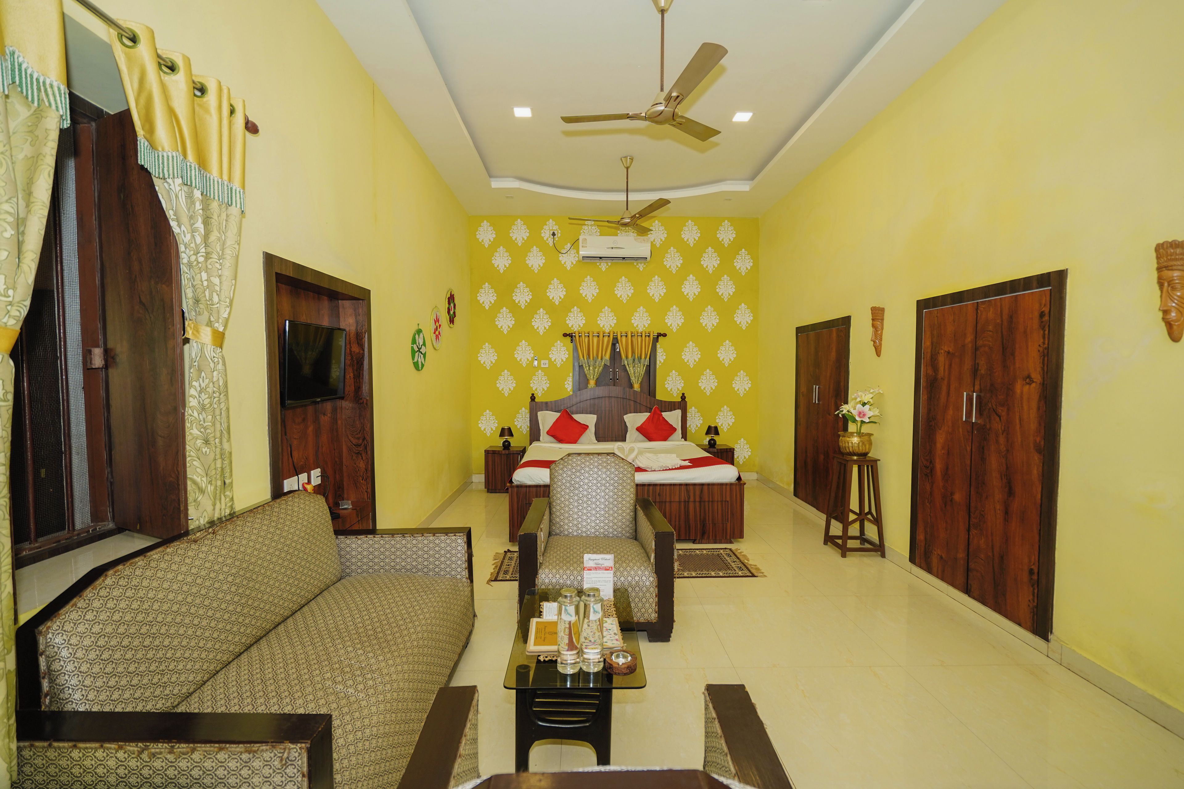 A hotel room with a sofa, a bed with red pillows, a ceiling fan, and a wall-mounted air conditioner.