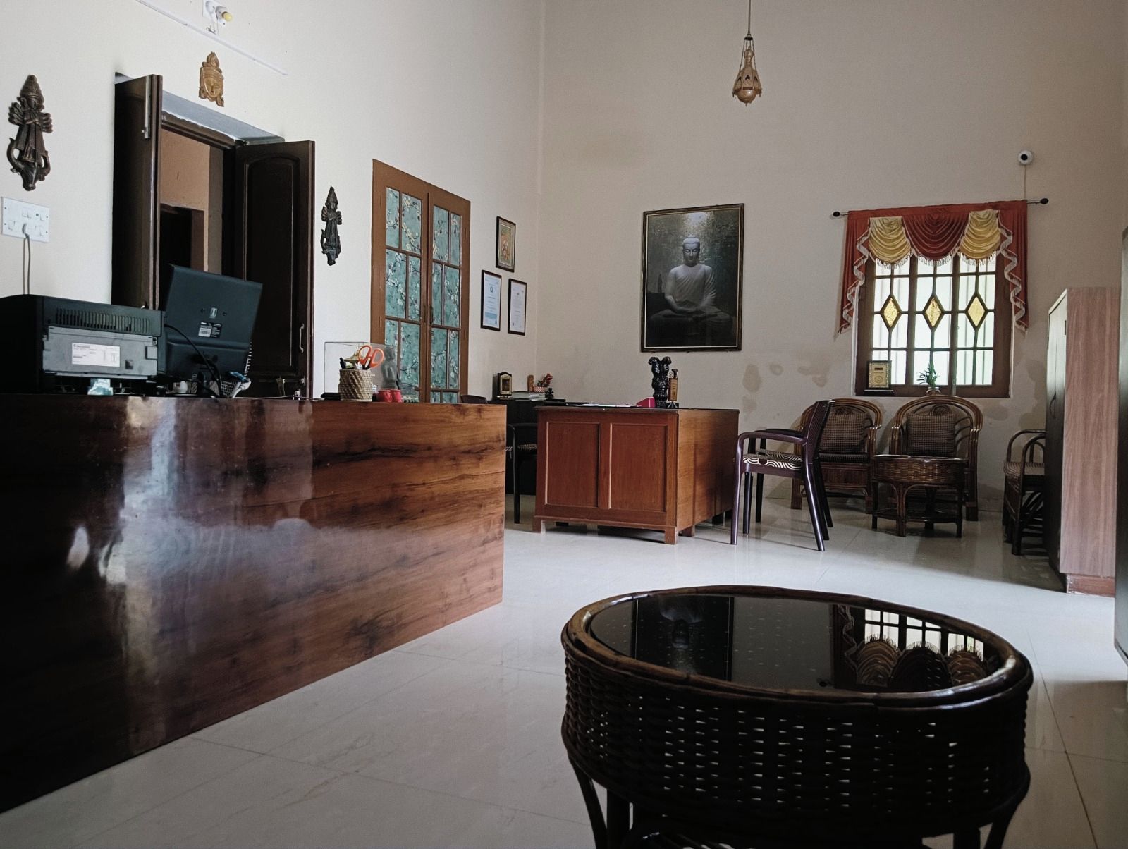Amplia habitación con un mostrador de recepción de madera, un gran ventanal con cortinas decorativas, una estatua de Buda montada en la pared y una silla de mimbre.