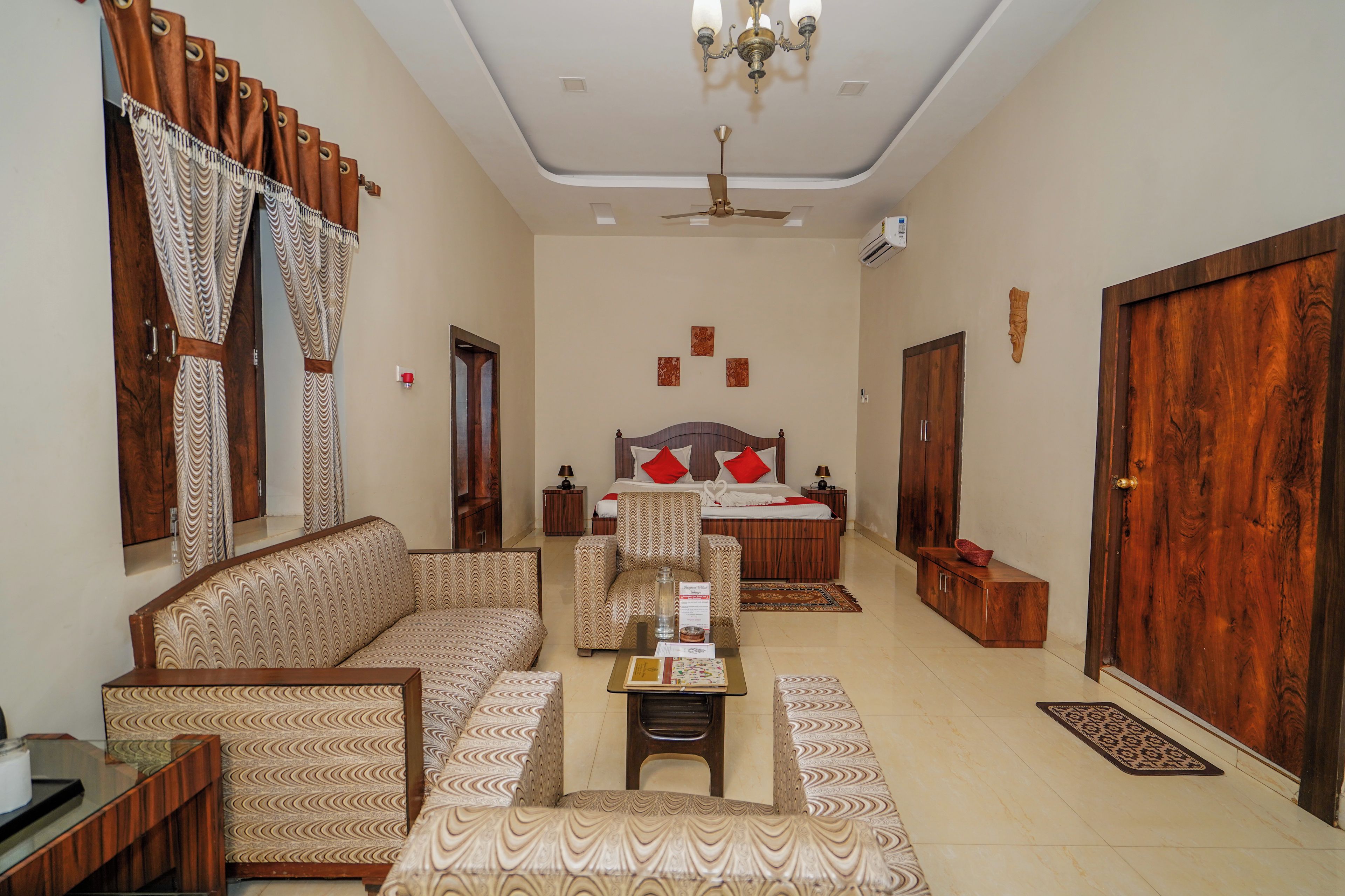 Una habitación de hotel con sofá, cama con almohadas rojas, armario de madera, candelabro y ventilador de techo.