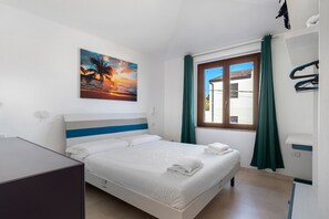 Ein Hotelzimmer mit einem Bett, einem Sonnenuntergangsgemälde, einem Fenster mit Vorhängen und Blick auf ein Gebäude.