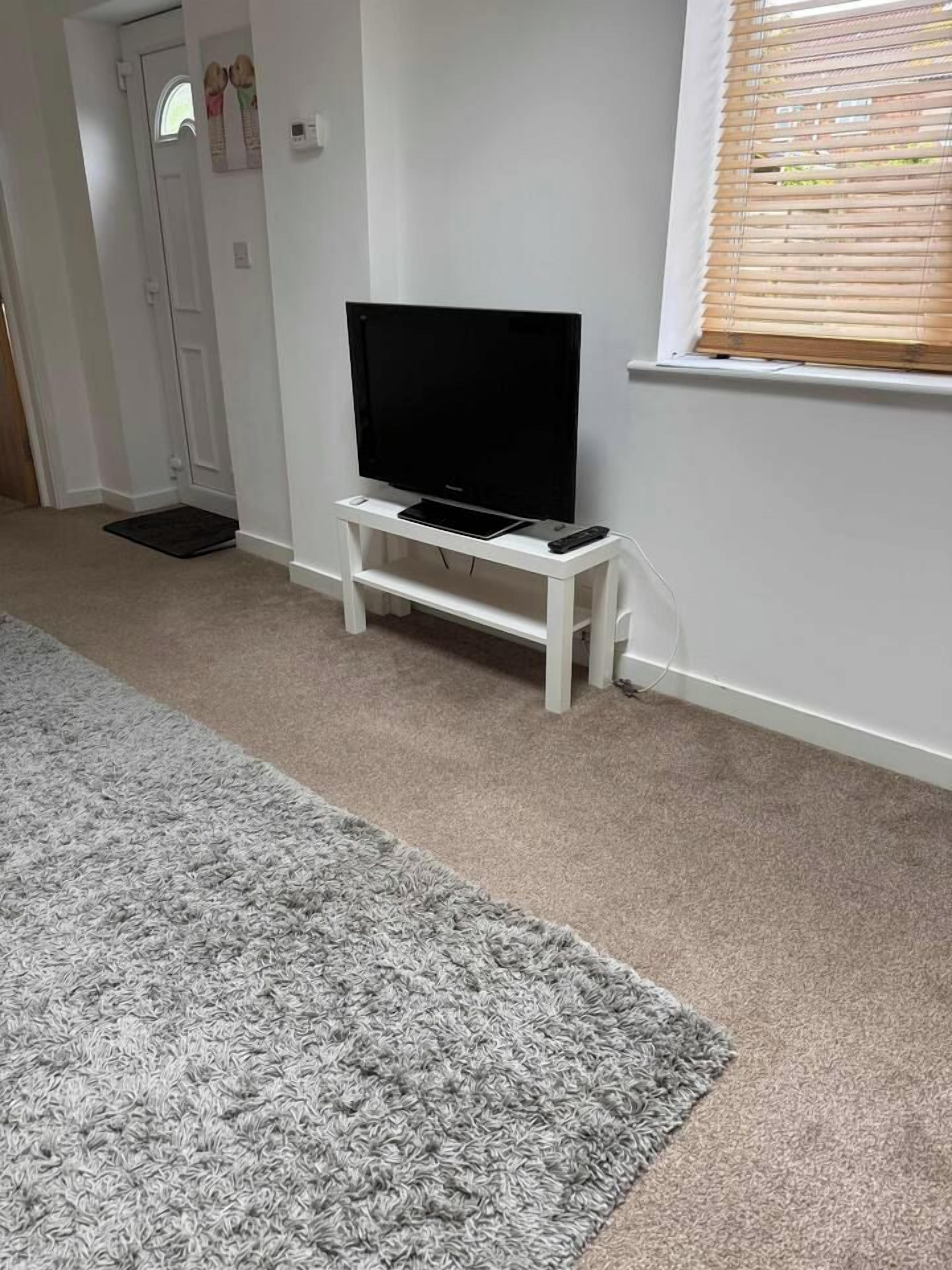 A room with a television on a white stand, a grey carpet, and a window with blinds.