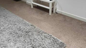 A room with a television on a white stand, a grey carpet, and a window with blinds.