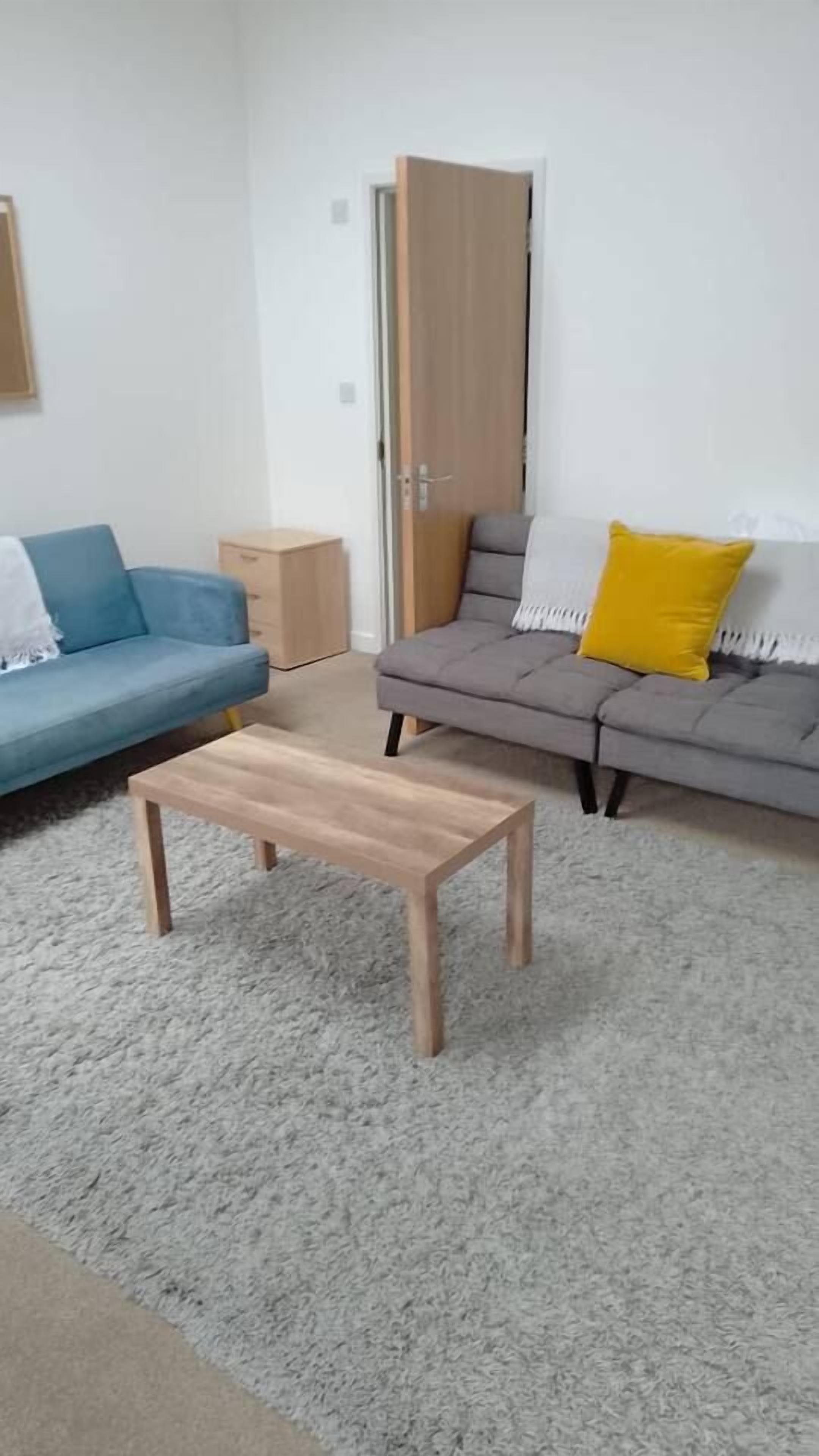 Een woonkamer met een blauwe bank, een grijze bank met een geel kussen en een houten salontafel.