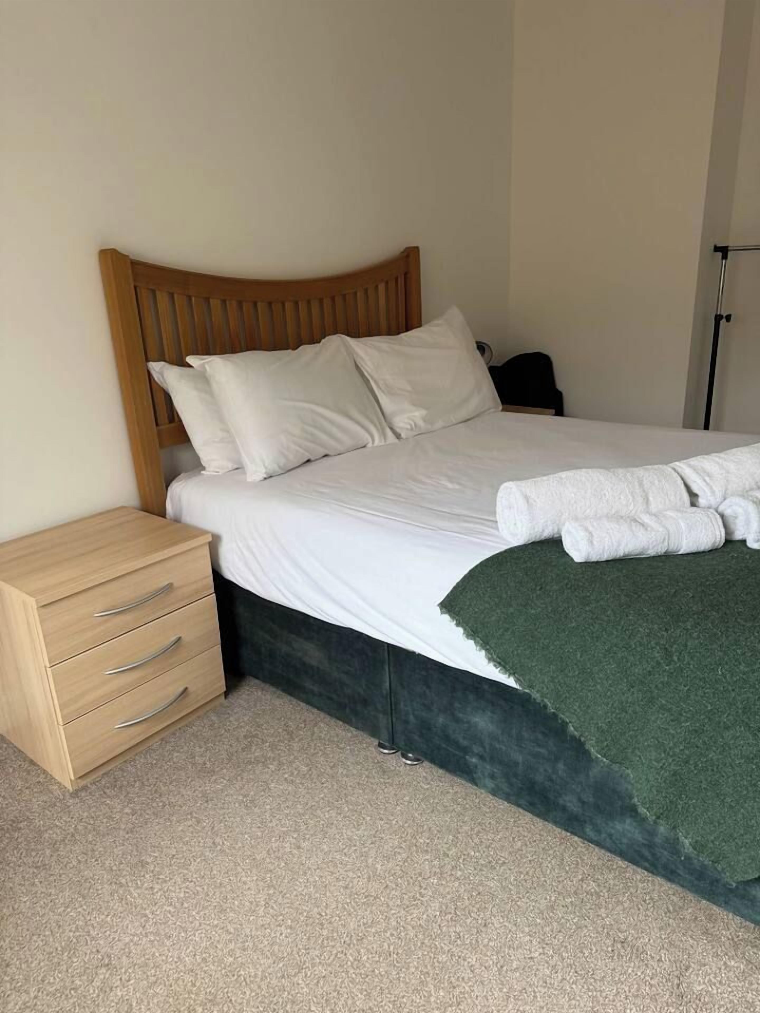 Een netjes opgemaakt bed met witte lakens, een houten hoofdbord en een groene plaid.