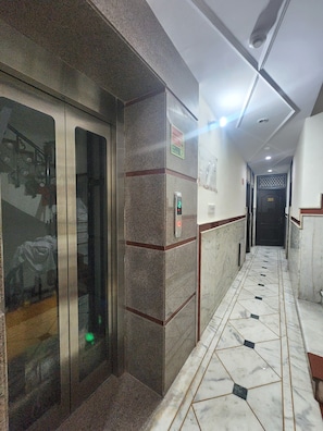 A hallway with a marble floor, a glass elevator, and a door marked with the number 305. - Oly Hotel Heritage Inn (New Delhi)