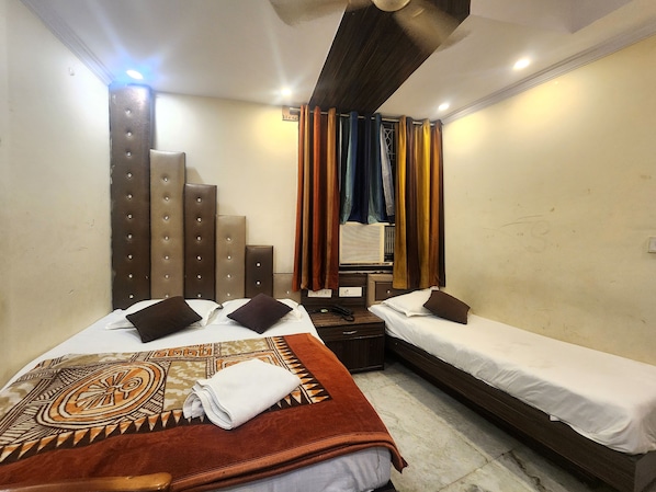 A hotel room with two beds, a large bed with a patterned blanket and pillows, and a smaller bed with a plain white bedspread. - Oly Hotel Heritage Inn (New Delhi)