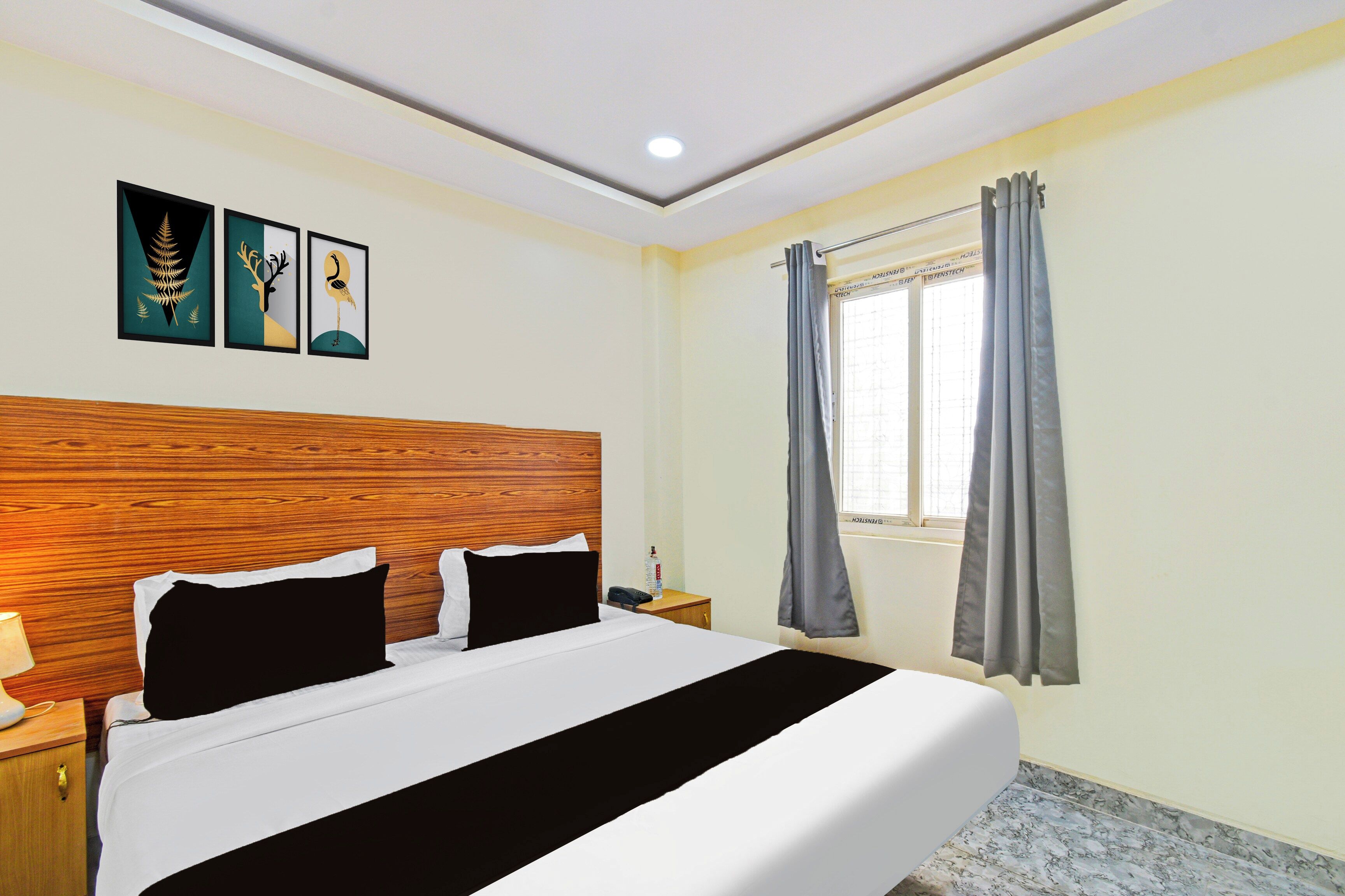 A hotel room with a bed, a nightstand, a lamp, a window with curtains, and two wall art pieces.