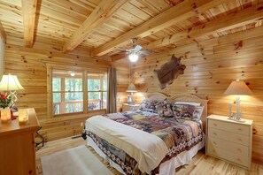 Un dormitori de cabana de fusta amb un llit, dues làmpades de nit, un ventilador de sostre, una finestra amb cortines i un pell de dibuix penjada.