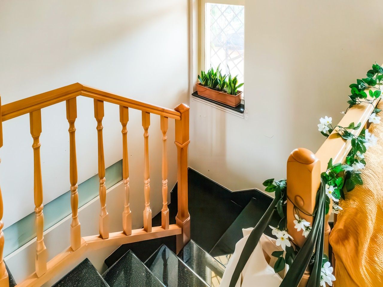 Un escalier avec des balustrades en bois, une fenêtre ornée d’une plante et une étagère murale décorée de fleurs.