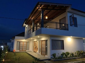 A modern two-story house with a balcony, large windows, and a well-lit exterior.