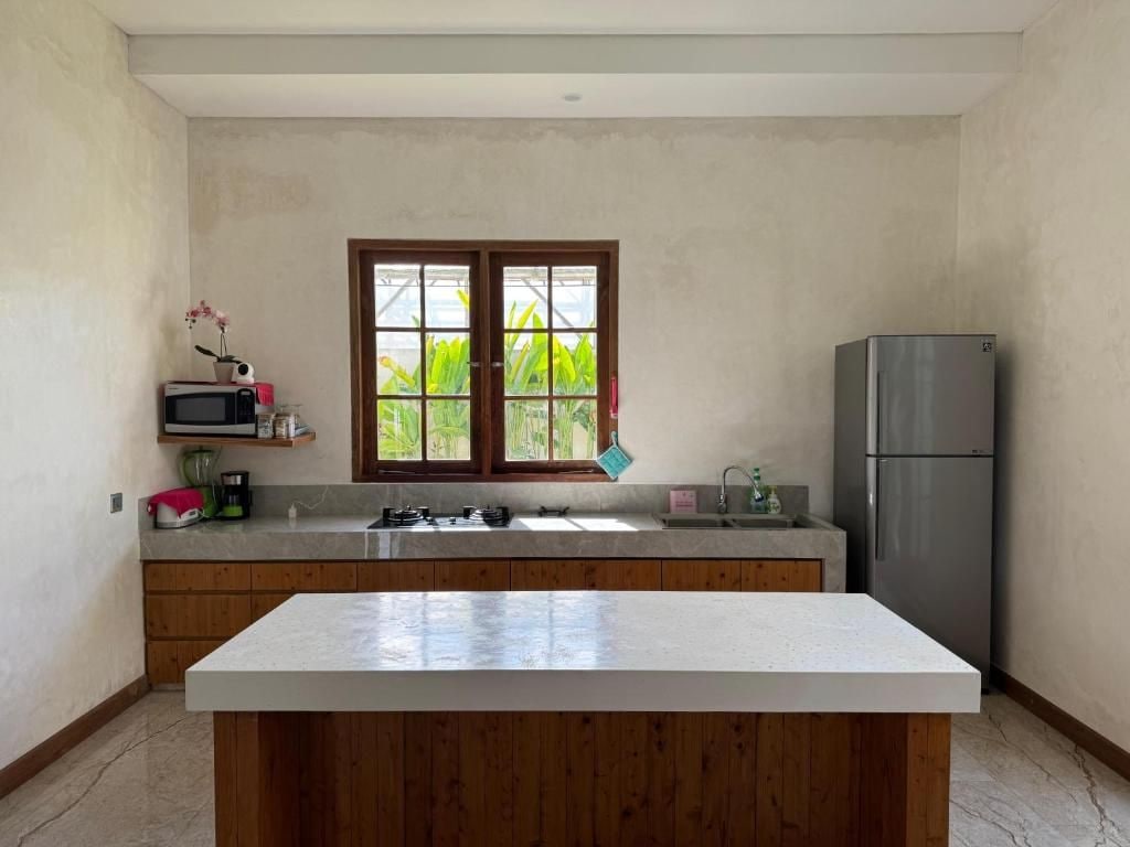 A kitchen with a central island, wooden cabinets, a stainless steel refrigerator, and a window with a view of greenery.