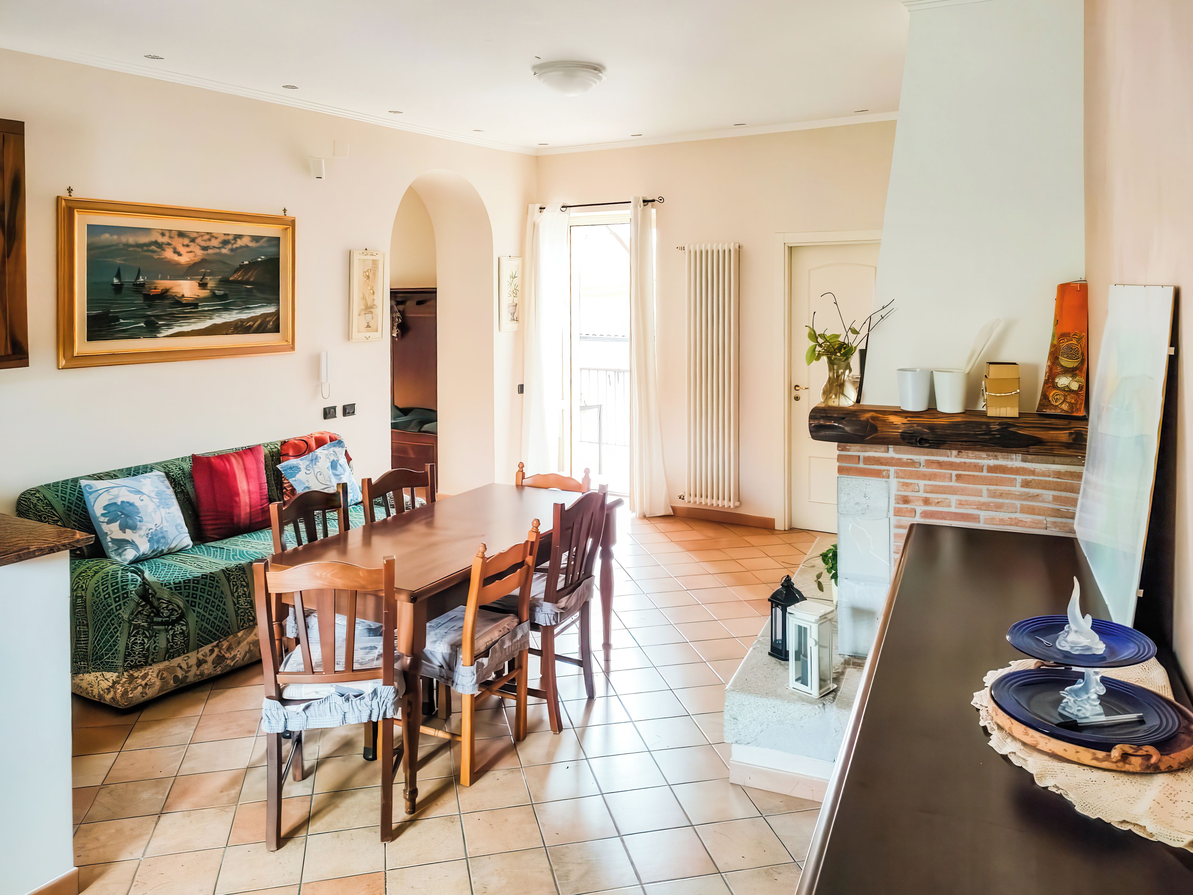 Un coin repas avec une table et des chaises en bois, un canapé vert agrémenté de coussins à motifs et une vue sur la mer par la fenêtre.