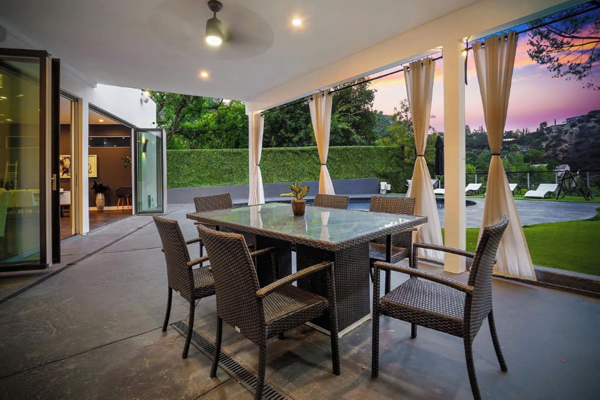 An outdoor patio with a glass-top table and wicker chairs, surrounded by curtains and a view of a garden.