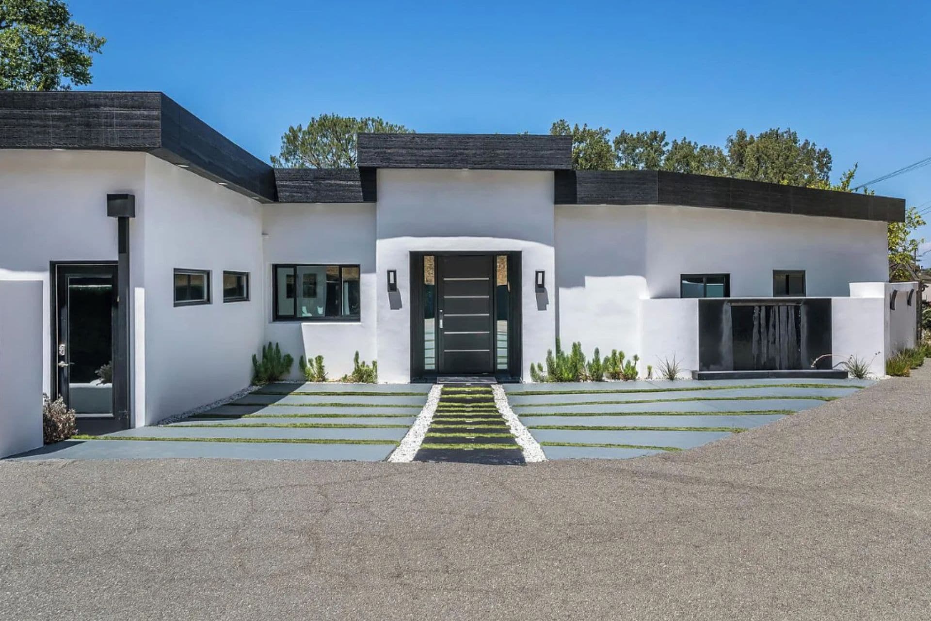 A modern two-story house with a flat roof, a front entrance with a black door, and a small garden area with a pathway leading to the entrance.