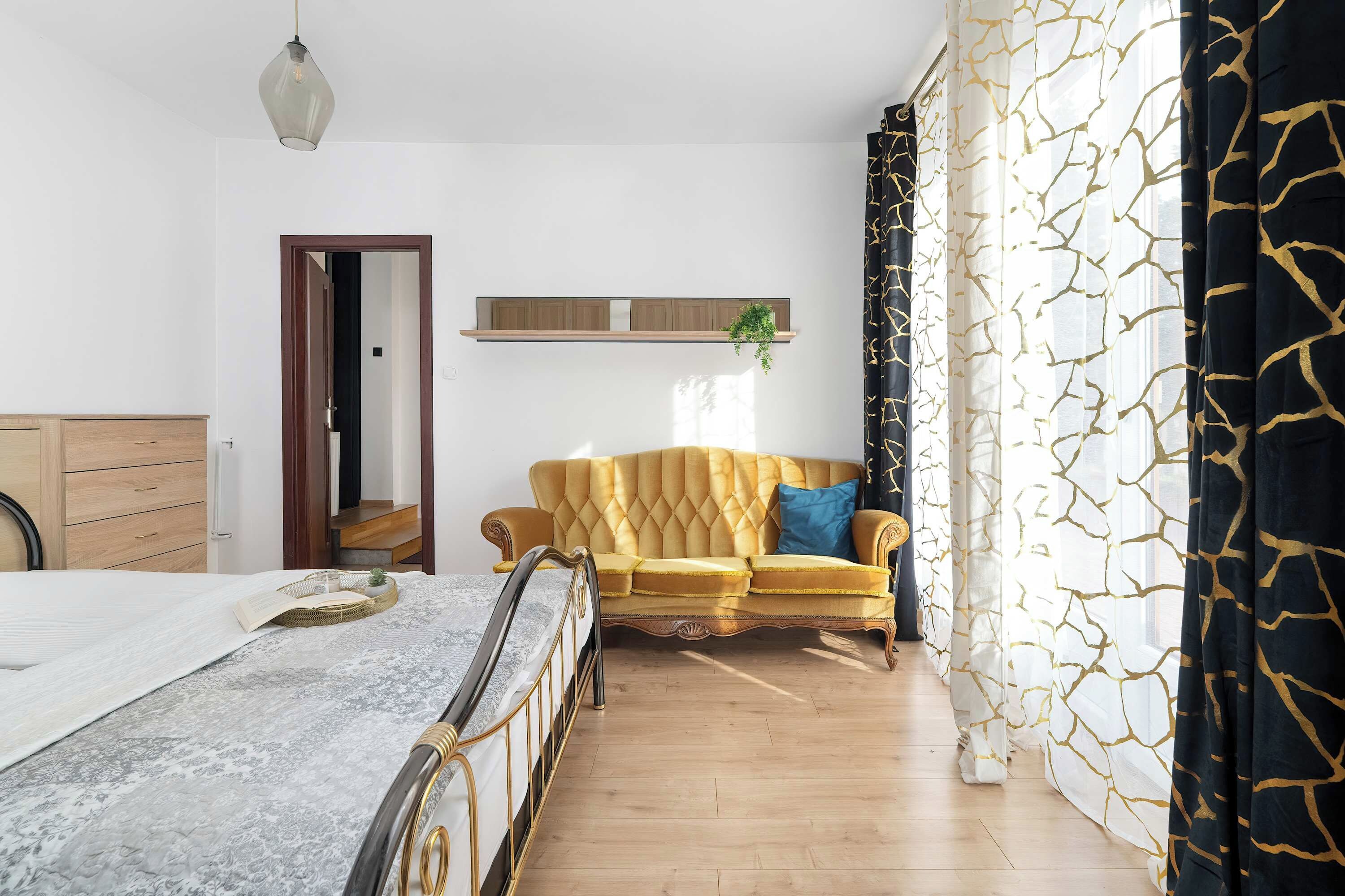 A bedroom with a bed, a sofa, a wooden dresser, a hanging plant, and a window with curtains.