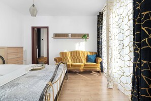 Une chambre à coucher comprenant un lit, un canapé, une commode en bois, une plante suspendue et une fenêtre avec des rideaux.