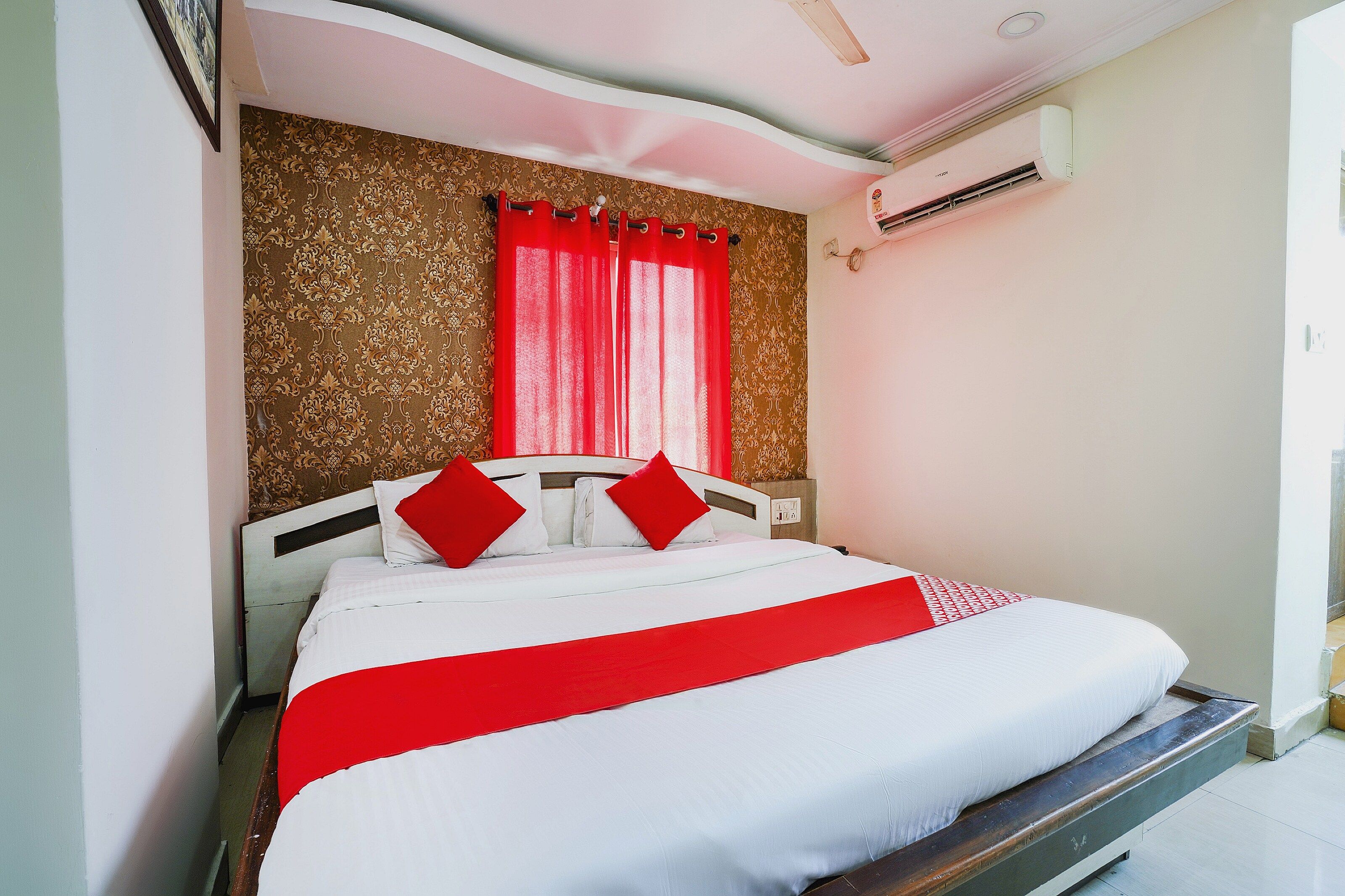 Cama com roupa de cama branca e detalhes vermelhos, parede estampada ao fundo e aparelho de ar condicionado na parede.