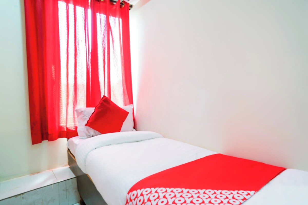 Una cama individual con ropa de cama blanca y una almohada roja decorativa, ubicada frente a una pared con un ventanal cubierto por cortinas rojas.