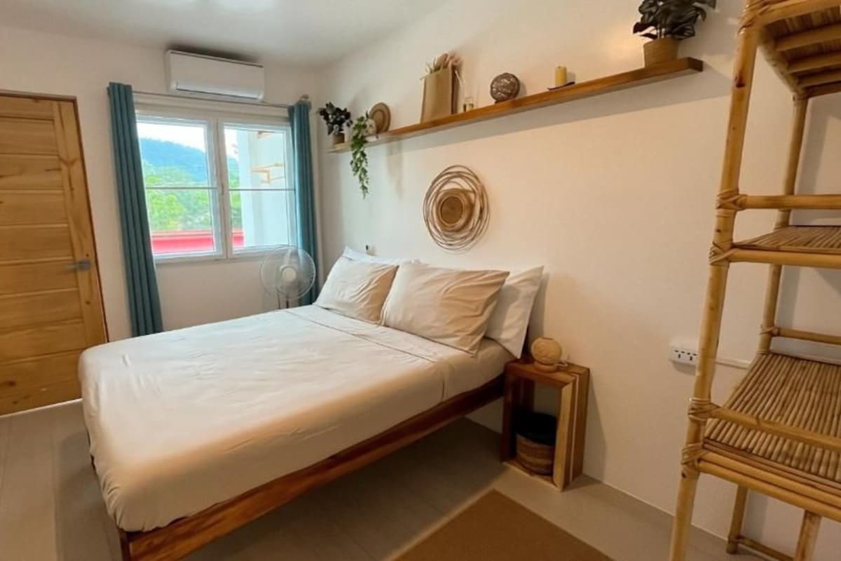 A bedroom with a bed, a wooden door, a window with curtains, and a wall-mounted shelf with decorative items.