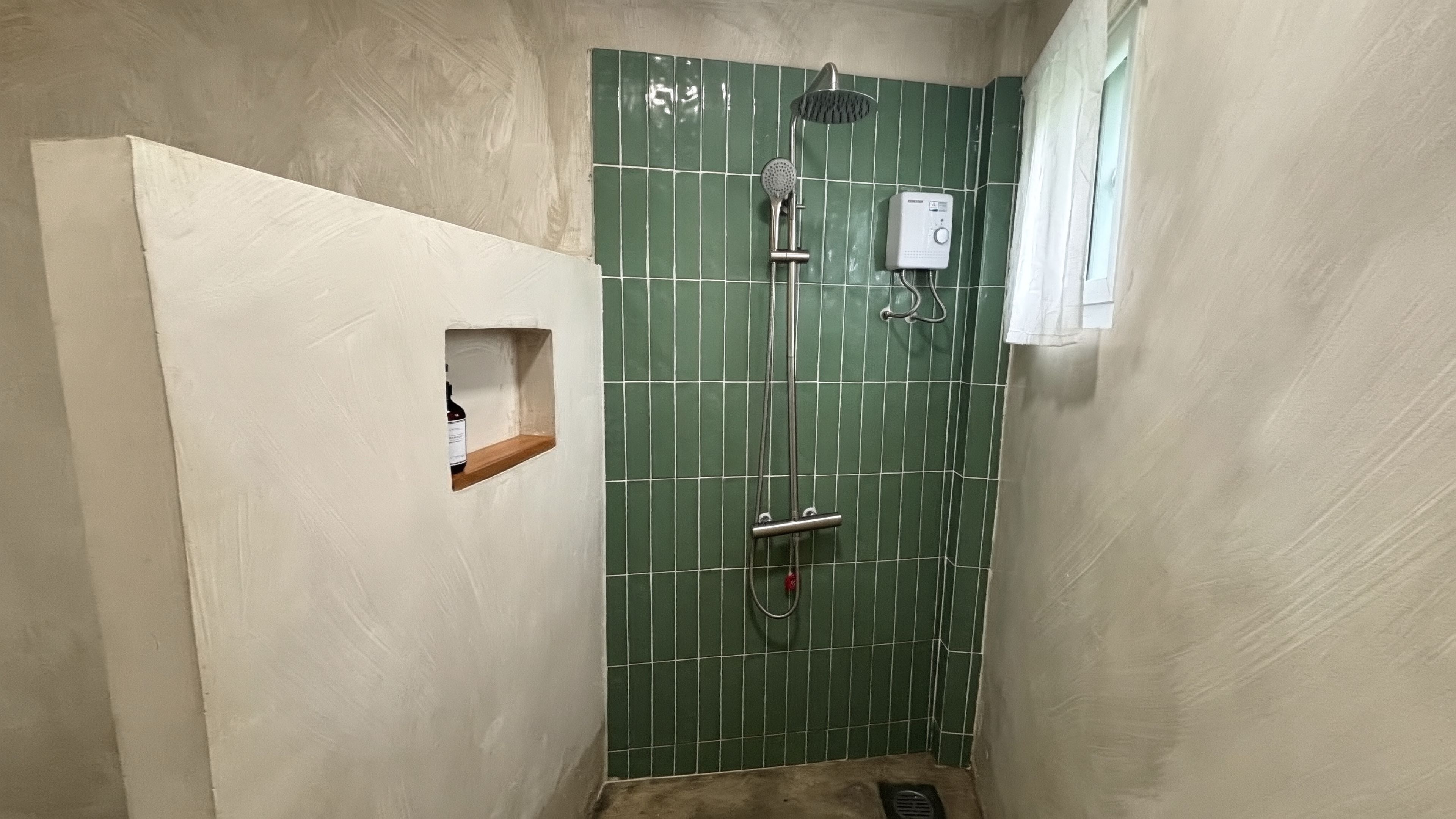 A small, narrow bathroom with a green-tiled shower wall, a handheld showerhead, a wall-mounted control panel, and a small shelf with a bottle.