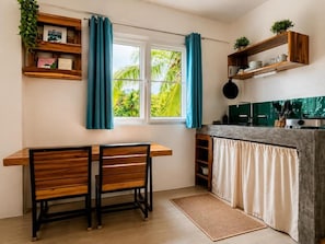 A kitchen with a dining table, chairs, a window with curtains, and shelves with kitchenware. - Hiraya Apartments (San Vicente)