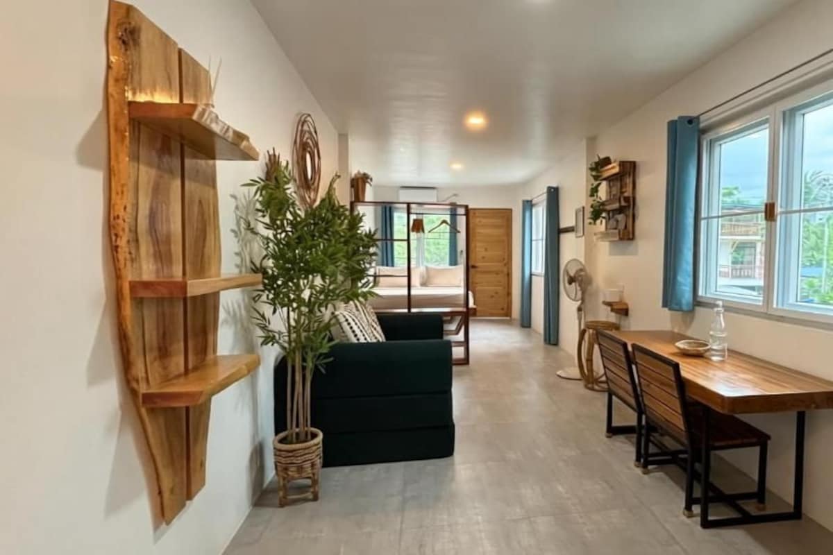 A room with a wooden shelf, a green sofa, a dining table with chairs, and a window with curtains.