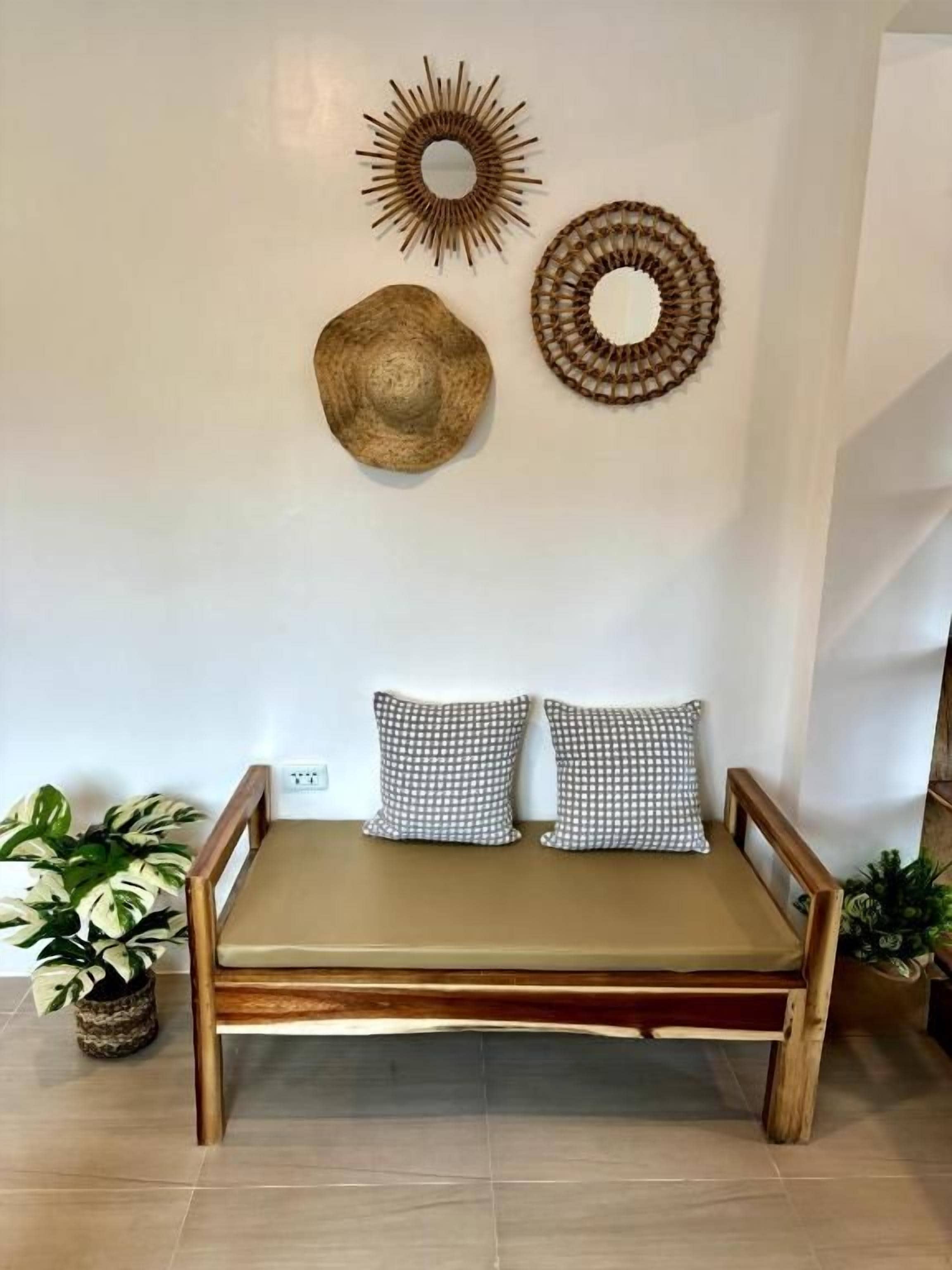 Ein Zimmer mit einer hölzernen Bank, zwei gemusterten Kissen und Wanddekoration, darunter ein Spiegel in Sonnenform und ein gewebter Korb.