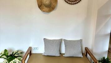 Habitación con un banco de madera, dos almohadas estampadas y decoración de pared que incluye un espejo con diseño de sol y una cesta tejida.