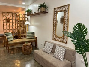 A room with a wooden bench, a cushioned sofa, a woven partition, a mirror, and a large plant.