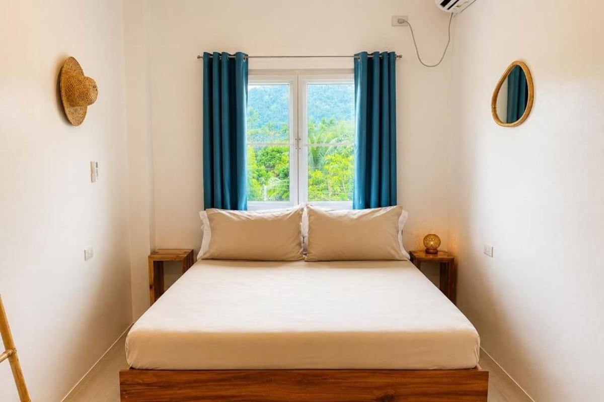 A room with a bed, a window with curtains, a wall-mounted mirror, and a wall-mounted hat.