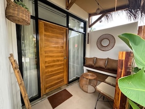 A room with a wooden door, a wicker chair, a round woven stool, and a bench with cushions.