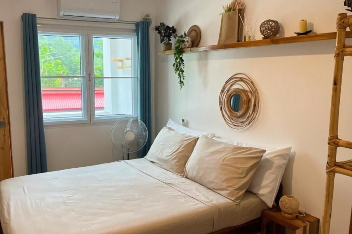 Una habitación con cama, ventana con cortinas, ventilador y repisa con objetos decorativos.