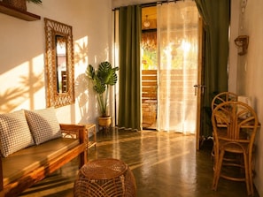 A room with a sofa, wicker chairs, a potted plant, and a sliding glass door leading to an outdoor area. - Hiraya Apartments (San Vicente)