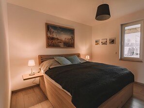 A bedroom with a bed, bedside lamps, a painting, and a window. - House 11 - Meadow Pipit (Am Salzhaff)