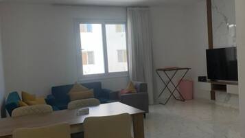 Een woonkamer met een eettafel, stoelen, een bank, een televisie en een raam met gordijnen.