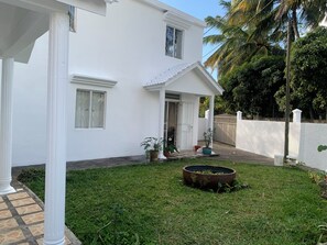 A white building with a covered porch, a small garden with a circular planter, and a paved walkway. - La Case de L'Artiste- 2 bedroom appartment in Bain Boeuf, 5 mins from the beach. (Cap Malheureux)