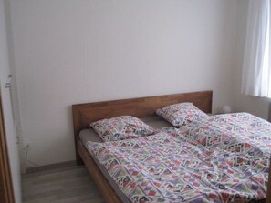 Cama de solteiro com colcha estampada e almofadas em quarto com paredes lisas e cabeceira de madeira.