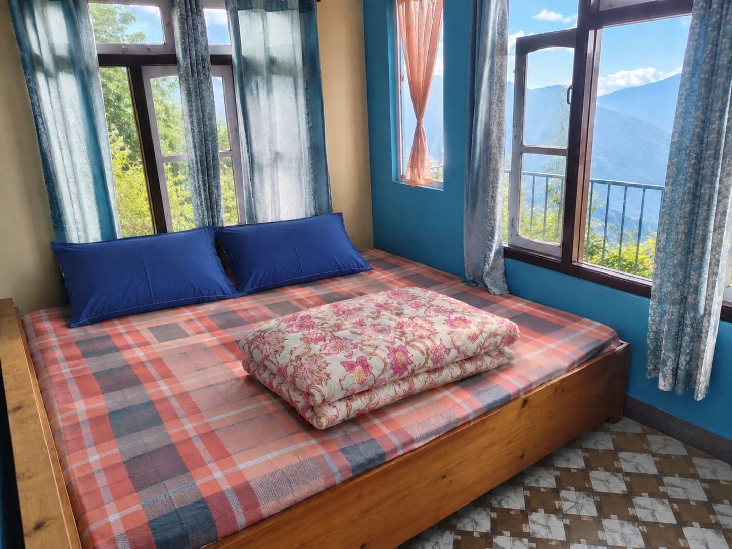 A bed with a plaid bedspread and a floral blanket, two blue pillows, and a view of mountains through the window.