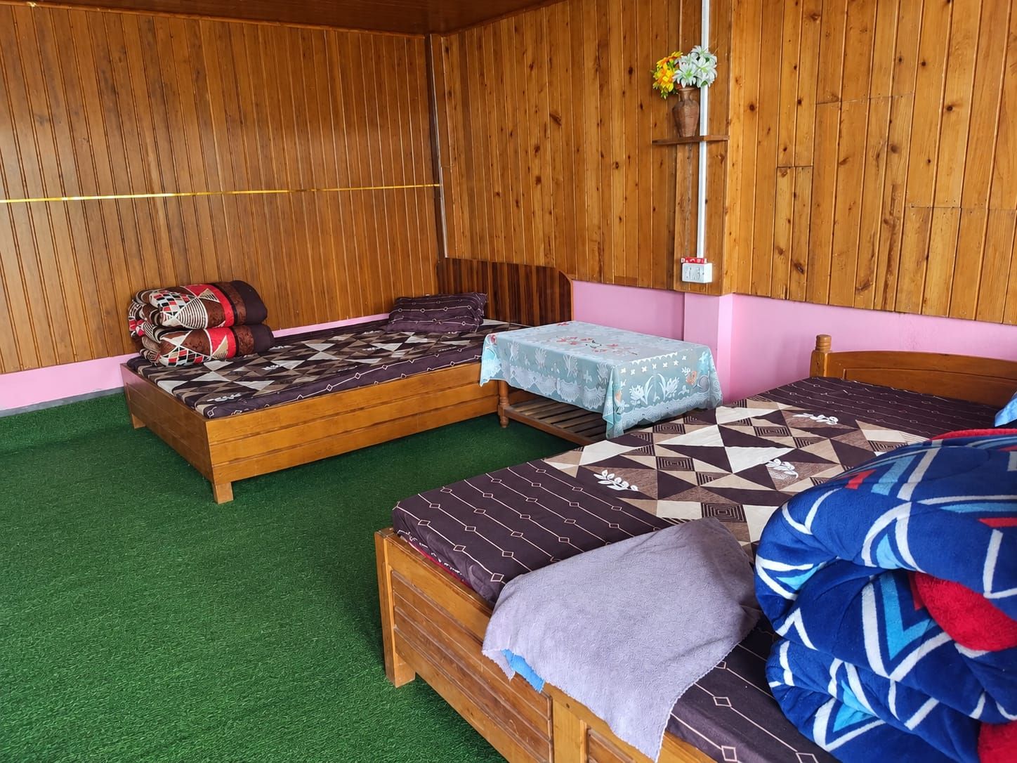 Una habitación con dos camas de madera, una mesita pequeña y una estantería con flores.