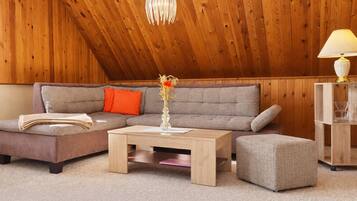 Ein gemütliches Zimmer mit Holzbalkendecke, einem grauen Sofa, einem Holztisch und einem kleinen Hocker.