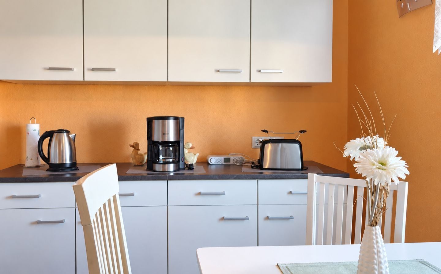 Una cocina con armarios blancos, cafetera, tostadora y un florero con flores en la encimera.