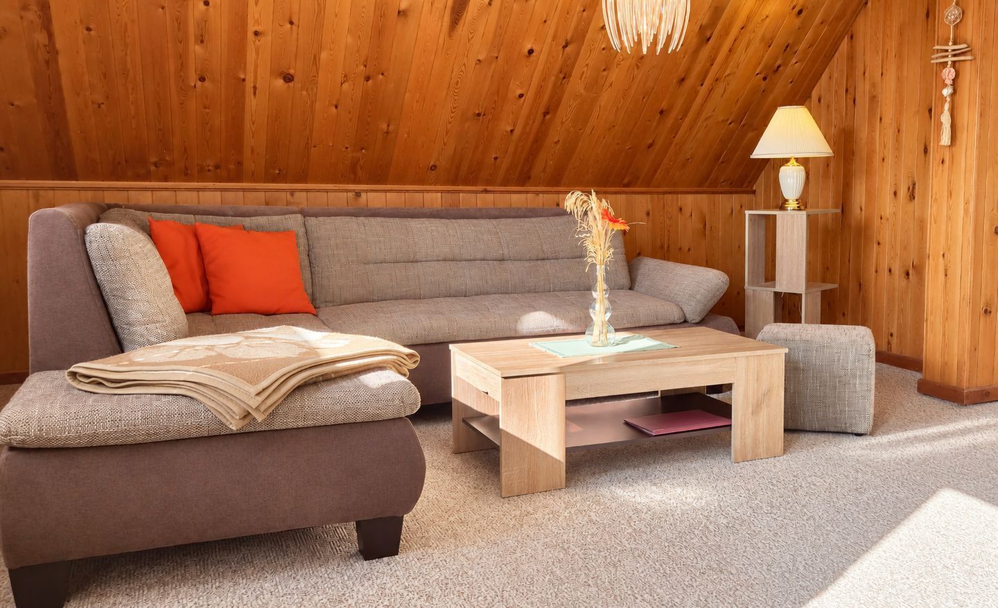 Een knus vertrek met een houten plafond, een grijze bank, een klein tafeltje en een lamp.