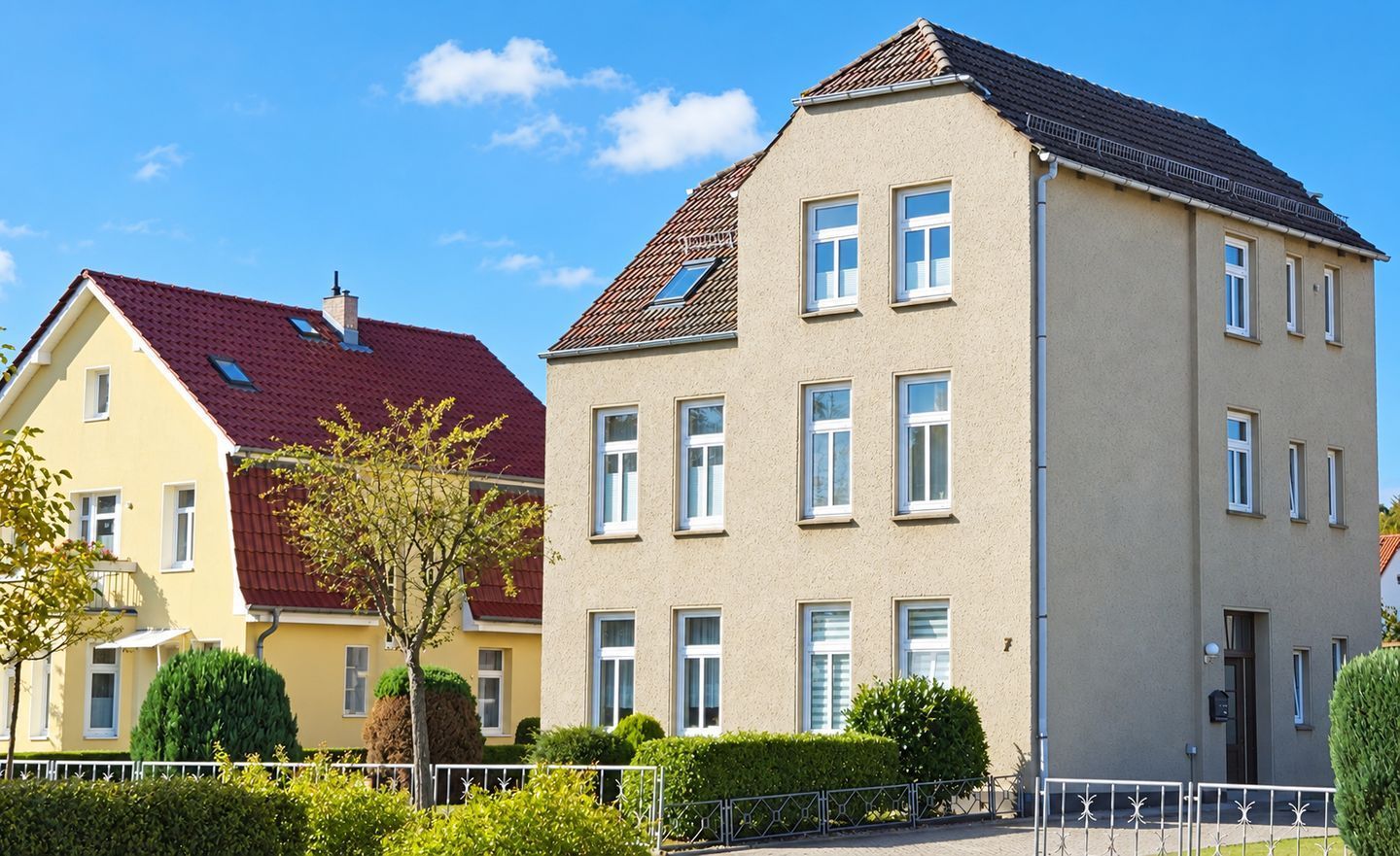 Ein zweistöckiges Wohngebäude mit rotem Ziegeldach, zahlreichen Fenstern und einem gepflegten Vorgarten mit Hecke und kleinem Baum.