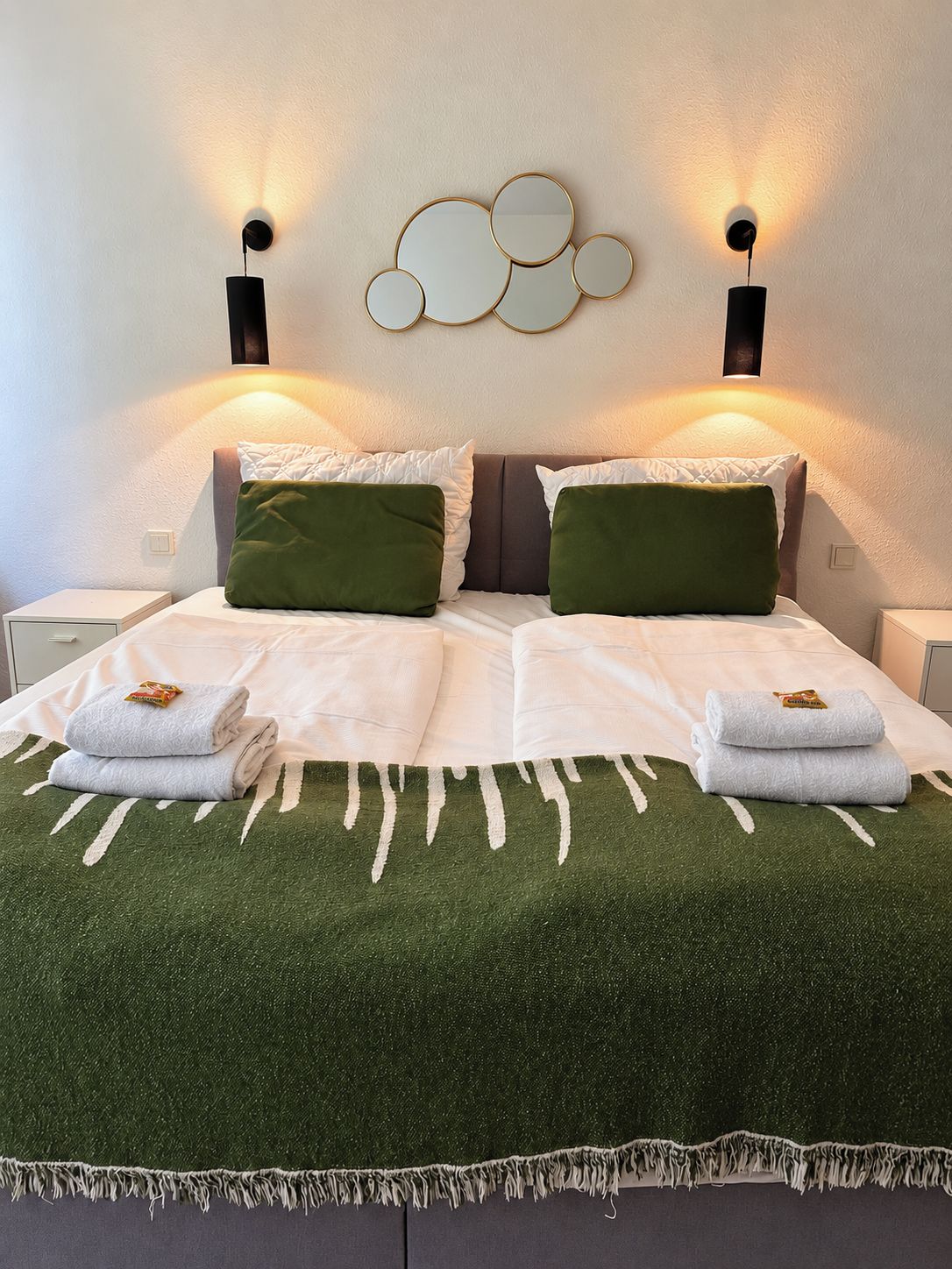 A bed with a green blanket, two pillows, and two folded towels. There are wall sconces with lights on either side of the bed.