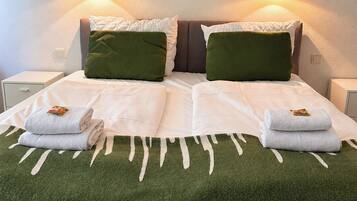 A bed with a green blanket, two pillows, and two folded towels. There are wall sconces with lights on either side of the bed.