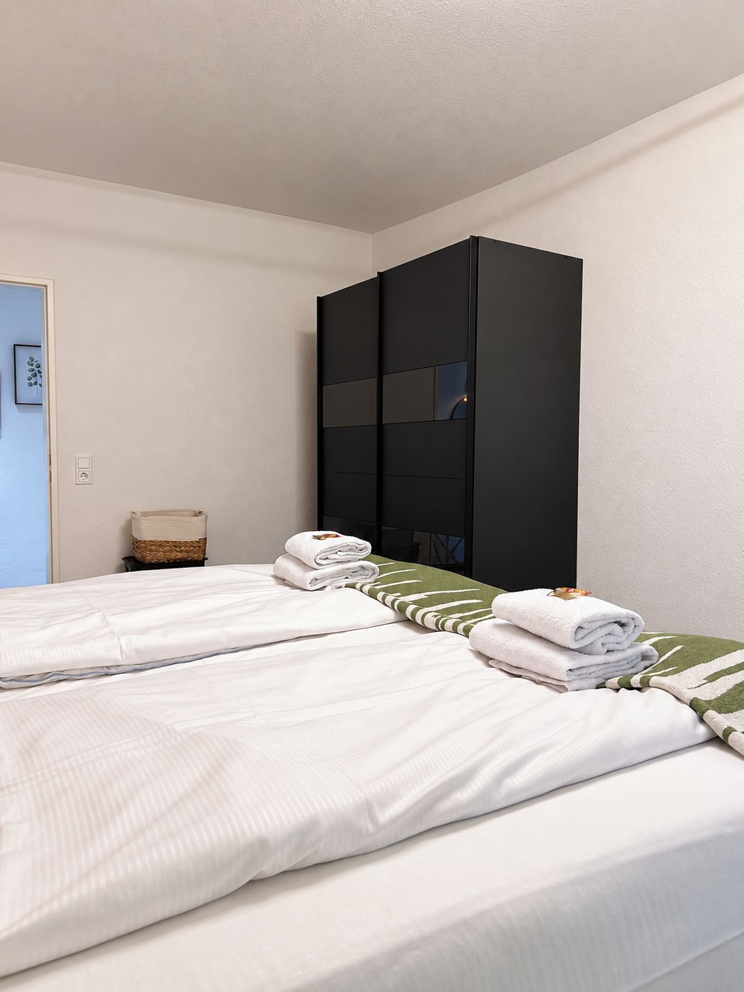 A hotel room with two beds, a wardrobe, a small table with a basket, and a framed picture on the wall.